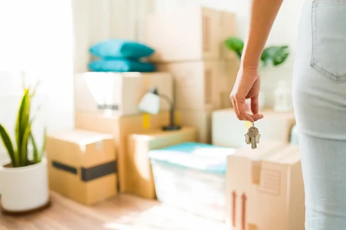 A woman standing in front of her moving boxes and plants in her new apartment, holding new keys.