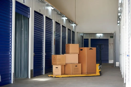 Stacked cardboard boxes on a pallet jack in a clean hallway lined with blue self storage unit doors.