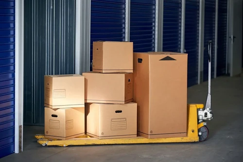 A stack of cardboard boxes on a yellow pallet jack inside a storage facility with blue roll-up doors.